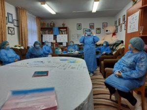 Six women in blue personal protective equipment (masks, hair coverings, gowns, gloves) sit socially distanced on chairs and couches around the perimeter of the room. One woman stands in the middle and demonstrates how to put on a mask.