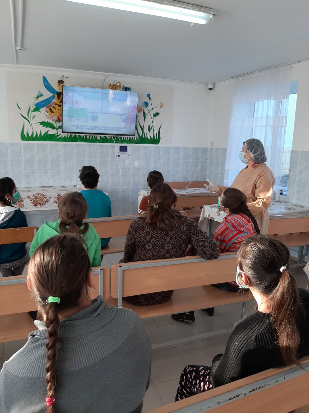 A masked audience sits on benches and watches a presentation on a large monitor on the wall.